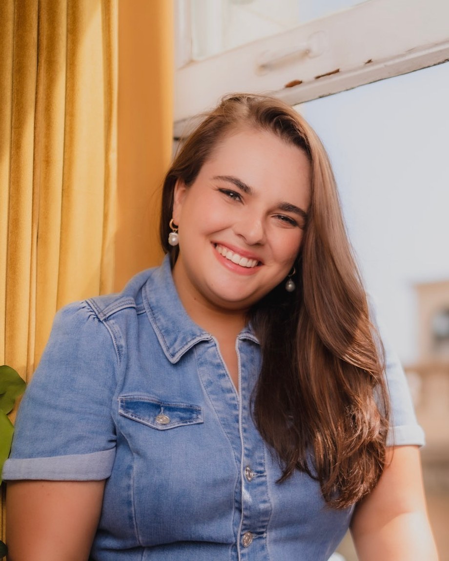 Andrea Lynn Piazza in a denim jumpsuit, seated by a window and smiling